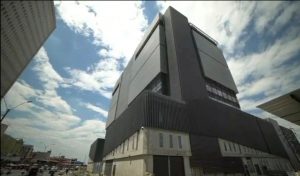 En la imagen se observa un edificio grande y moderno con una arquitectura robusta y de líneas rectas. La fachada está compuesta principalmente por paneles metálicos de color oscuro y secciones de concreto claro en la parte inferior. La estructura tiene un aspecto industrial o tecnológico, con pocas ventanas visibles y una apariencia sólida y cerrada.El cielo está parcialmente nublado, con zonas de azul visible entre las nubes, lo que da buena iluminación a la escena. En la parte baja de la imagen se ven vehículos y motocicletas circulando por la calle, además de algunos edificios cercanos y postes de luz, lo que sugiere que se trata de una zona urbana.