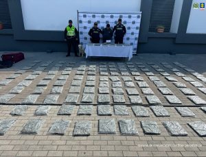 En la imagen se observa un operativo de las autoridades. Cuatro uniformados, junto con un perro guía, posan detrás de una mesa donde reposan varios paquetes incautados. Frente a ellos, sobre el suelo, hay decenas de bultos o paquetes rectangulares envueltos en material plástico, dispuestos en filas ordenadas. Detrás del grupo se aprecia un pendón institucional con los logotipos oficiales de la Policía Nacional y la Fiscalía General de la Nación, lo que sugiere que se trata de una presentación de resultados de un operativo contra el tráfico de estupefacientes.