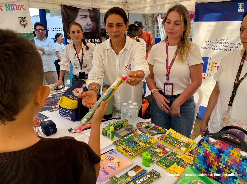 En la imagen aparecen tres funcionarias de la Fiscalía, entregando un lápiz a un niño, frente a una mesa llena de material didáctico y de prevención.