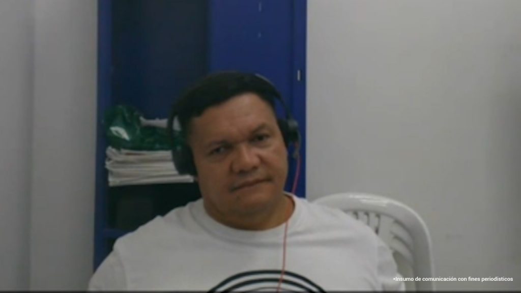 En la imagen aparece un hombre de contextura robusta, con cabello corto y oscuro, sentado en una silla plástica blanca. Lleva puesta una camiseta blanca y unos audífonos negros con cable rojo. Detrás de él se observan una pared blanca y un estante azul con algunos objetos. Su expresión es seria y parece estar participando en una audiencia virtual.