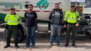 En la imagen se ven dos personas capturadas que visten ropa informal y cada costado dos policías. De fondo se ve un camión tipo cisterna.