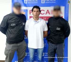 Tres hombres están frente a un fondo azul con logos de la Fiscalía. En el centro hay un hombre joven, con camiseta blanca y las manos hacia atrás. A cada lado hay funcionarios del CTI y de la Policía Judicial, vestidos con chaquetas negras y el rostro difuminado.