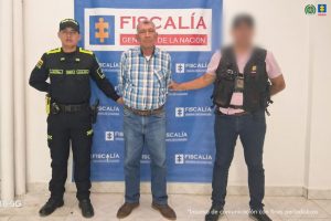 En la imagen se ve a tres personas posando frente a un fondo institucional. Esto es lo que se observa:En el centro hay un hombre mayor, vestido con una camisa de cuadros y jeans, con las manos atrás como si estuviera siendo custodiado.  A cada lado hay agentes uniformados:  A la izquierda, un policía con uniforme negro, chaleco reflectivo y equipo táctico.  A la derecha, un agente con chaleco táctico, cuya cara está difuminada, posiblemente para proteger su identidad.  Detrás de ellos hay un telón azul con logotipos que dicen “FISCALÍA GENERAL DE LA NACIÓN”, lo que sugiere que la fotografía es parte de un registro oficial o comunicado institucional.  En la esquina superior derecha aparecen emblemas oficiales.