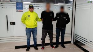 La imagen muestra a tres personas de pie en un espacio cerrado con paredes de vidrio y puertas corredizas. Dos de ellas llevan chaquetas institucionales:Persona a la izquierda: viste una chaqueta amarilla con el logotipo de la Policía y jeans azules.
Persona a la derecha: lleva una chaqueta negra con las siglas CTI y el texto “Policía Judicial” visibles en la parte frontal.
Persona en el centro: está vestida con camiseta negra con el logo de Jordan y pantalón oscuro; tiene grilletes en los tobillos. En el fondo se observa un letrero que dice:
“Grupo Técnico Operativo Captura – Fiscalía”, indicando que el lugar corresponde a una dependencia de la Fiscalía. También se aprecia el escudo institucional en la parte superior derecha de la imagen y una nota en la esquina inferior que dice:
“Insumo de comunicación con fines periodísticos”.