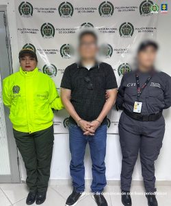 La imagen muestra a tres personas de pie frente a un fondo institucional que tiene repetidos los logotipos de la Policía Nacional de Colombia. El fondo es blanco con los escudos en color verde y negro.Persona a la izquierda: viste un uniforme verde fosforescente con el texto visible “SIJIN” y un código “OS-402” en la parte frontal.
Persona en el centro: lleva puesta una camiseta negra con puntos blancos, pantalón de mezclilla azul y zapatos oscuros. Sus manos están juntas al frente, sujetadas por las personas a los lados.
Persona a la derecha: viste un uniforme gris oscuro con el texto “POLICÍA JUDICIAL” en el pecho y “CTI” en la parte superior. También porta un cinturón táctico y una credencial colgada al cuello. En la esquina superior derecha del fondo aparece el logotipo de la Fiscalía General de la Nación. En la parte inferior de la imagen se lee la frase: “Insumo de comunicación con fines periodísticos”.