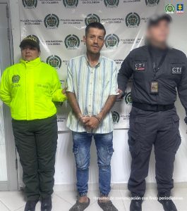 La foto muestra a tres personas de pie frente a un fondo institucional que tiene repetidamente el logotipo de la Policía Nacional de Colombia.Persona a la izquierda: viste un uniforme verde fosforescente con el texto “SIJIN” y un código “OS-402” en la parte frontal.
Persona en el centro: lleva una camiseta negra con pequeños puntos blancos, pantalón de mezclilla azul y zapatos oscuros. Sus manos están juntas al frente, sujetadas por las personas a los lados.
Persona a la derecha: viste un uniforme gris oscuro con el texto “POLICÍA JUDICIAL” en el pecho y “CTI” en la parte superior. Porta un cinturón táctico y una credencial colgada al cuello. En la esquina superior derecha del fondo aparece el logotipo de la Fiscalía General de la Nación. En la parte inferior de la imagen se lee la frase: “Insumo de comunicación con fines periodísticos”.