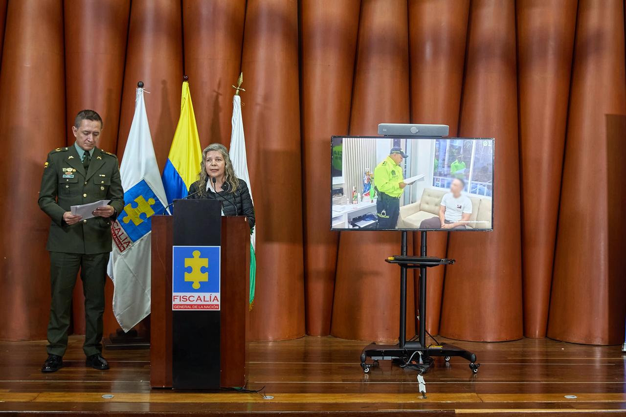 En la imagen se ve una rueda de prensa de la Fiscalía General de la Nación. Al centro, la delegada para las Finanzas Criminales, Aura Liliana Trujillo, está de pie detrás de un atril mientras ofrece declaraciones. A su lado izquierdo está un oficial de la Policía Nacional leyendo unos documentos. Detrás de ellos se observan tres banderas: la de la Fiscalía, la de Colombia y otra institucional. A la derecha de la escena hay una pantalla que muestra una fotografía del momento en que un uniformado notifica a uno de los capturados dentro de una vivienda. Todo ocurre en un salón con paredes de color marrón y piso de madera.
