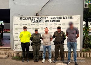 En la foto aparecen cinco personas posando frente a un telón institucional de la Policía Nacional de Colombia, específicamente de la Seccional de Tránsito y Transporte Bolívar – Cuadrante Vial Gambote.Descripción de la escena: Hay cuatro personas uniformadas, tres con uniformes de policía y una con una chaqueta amarilla reflectante. En el centro está un hombre sin uniforme, con las manos hacia atrás, aparentemente bajo custodia. A la derecha hay otra persona de pie con camiseta gris; su rostro está difuminado. Todos están alineados formando una fila para una fotografía oficial. El fondo muestra logos institucionales y textos de la policía.