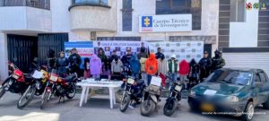 En la imagen se ve lo siguiente:Un grupo numeroso de personas alineadas frente a un edificio institucional. Varias de ellas tienen capuchas o ropa que cubre parcialmente sus rostros; otras llevan uniforme de fuerzas de seguridad. Detrás del grupo hay varios avisos y pendones oficiales, entre ellos uno del Cuerpo Técnico de Investigación y otro del GAULA Militar Nariño, lo que indica que se trata de un operativo conjunto de autoridades. Al frente hay varias motocicletas y un automóvil estacionados, algunos con papeles adheridos al frente (posiblemente como evidencia). También se observa una mesa con objetos decomisados, que parecen ser documentos, teléfonos y otros elementos. En general, la escena corresponde a una presentación pública de un operativo policial o militar, con personas retenidas y evidencia incautada.