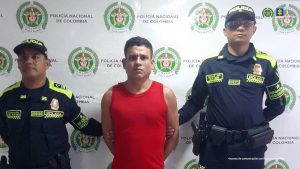 En la imagen se ve a tres personas de pie frente a un fondo institucional de la Policía Nacional de Colombia.A los costados hay dos uniformados con distintivos y chalecos oscuros con detalles en amarillo. Ambos llevan gorras de policía y tienen una postura firme. En el centro está un hombre con camiseta roja sin mangas, con las manos hacia atrás, sostenidas por los policías. Su expresión es seria y se encuentra claramente bajo custodia. En la parte superior derecha aparecen los logos de la Policía Nacional y de la Fiscalía.
