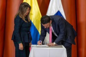En la imagen aparecen dos personas en un ambiente formal, posiblemente durante un acto oficial o ceremonia.A la izquierda, hay una mujer vestida con un traje oscuro con detalles verdes. Está de pie, observando atentamente. A la derecha, hay un hombre con traje oscuro y corbata rosada que se inclina hacia adelante para firmar un documento sobre una mesa cubierta con un mantel blanco. Detrás de ellos se ven dos banderas: una con los colores amarillo, azul y rojo, y otra blanca con un diseño en azul. El entorno sugiere un evento protocolario, como la firma de un acuerdo, posesión de un cargo o un acto gubernamental.