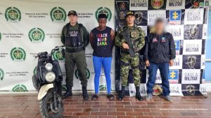 Al centro hay una persona detenida, de pie, con la cabeza ligeramente inclinada hacia abajo y las manos al frente. Viste una camiseta sin mangas oscura con un texto multicolor y pantalón azul.A ambos lados de esta persona hay miembros de fuerzas de seguridad: algunos con uniformes militares camuflados y otros con chalecos y ropa identificada con insignias oficiales.  Detrás de ellos se ve un fondo con logotipos repetidos que incluyen “Policía Nacional de Colombia”, “GAULA Militar”, “Fuerzas Militares” y “Fiscalía”, lo que sugiere un contexto de captura o procedimiento judicial.  A la izquierda de la imagen hay una motocicleta exhibida, aparentemente como elemento incautado.  Uno de los agentes sostiene un arma larga, y otro tiene el rostro intencionalmente desenfocado, posiblemente por motivos de seguridad.  En la parte inferior derecha aparece un texto que indica que es un insumo de comunicación con fines periodísticos.  En conjunto, la imagen parece documentar una detención oficial y la presentación de una persona capturada junto a autoridades colombianas.