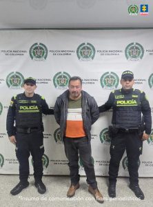 En la foto se observa a tres personas posando frente a un fondo institucional de la Policía Nacional de Colombia.Al centro hay un hombre con ropa civil (chaqueta oscura y camiseta de varios colores), con las manos hacia atrás, en una postura que sugiere que está bajo custodia. A ambos lados del hombre hay dos agentes de policía uniformados, con chalecos y gorras que dicen “POLICÍA”. Cada uno lo sostiene ligeramente por un brazo. El fondo repite el emblema y el texto de la Policía Nacional, típico de fotografías oficiales o comunicados. En la parte inferior de la imagen aparece el texto: “Insumo de comunicación con fines periodísticos”, lo que indica que la foto está destinada a difusión informativa.