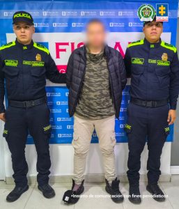 En la imagen se observan tres personas de pie frente a un fondo institucional azul con el logotipo y la palabra “Fiscalía” repetidos.En el centro hay una persona con el rostro intencionalmente desenfocado, vestida con una chaqueta acolchada oscura, una camiseta tipo camuflaje, pantalón claro y zapatos oscuros. Sus manos parecen estar hacia atrás, y está flanqueada por las otras dos personas.  A ambos lados hay dos agentes de policía con uniforme azul oscuro y detalles verde neón, gorras que dicen “Policía”, botas negras y parches identificativos en el uniforme. Cada uno sostiene al individuo del centro por un brazo.  En la parte superior derecha del fondo aparecen emblemas oficiales, y en la parte inferior de la imagen se lee el texto:
“Insumo de comunicación con fines periodísticos”.