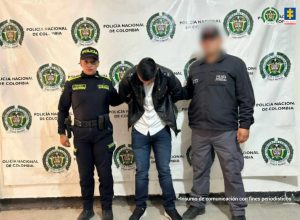 En la imagen se observa a tres personas frente a un fondo institucional con los logotipos de la Policía Nacional de Colombia y la Fiscalía.En el centro, hay un hombre con la cabeza inclinada hacia abajo, vestido con una chaqueta negra, camisa blanca y jeans. Sus brazos están sujetos por las personas a ambos lados. A la izquierda, aparece una agente de la Policía Nacional, con uniforme oscuro, gorra y cinturón de dotación. Su postura es firme y está mirando hacia la cámara. A la derecha, se ve otra persona con chaqueta oscura y pantalón gris, cuyo rostro está difuminado, lo que sugiere protección de identidad. En su vestimenta se aprecia un distintivo relacionado con funciones judiciales.