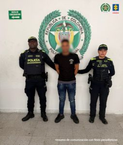 Las dos personas ubicadas a los lados visten uniformes oscuros de la Policía Nacional de Colombia, con chalecos tácticos, insignias visibles y equipos de dotación. Se mantienen erguidas y con postura formal.La persona ubicada en el centro viste ropa de civil: una camiseta oscura, jeans azules y calzado deportivo. Tiene los brazos hacia atrás y está flanqueada por los uniformados. El piso es de baldosa clara y el entorno corresponde a un espacio institucional cerrado, probablemente dentro de una sede oficial. En la parte inferior de la imagen se incluye el texto: “Insumo de comunicación con fines periodísticos”,
