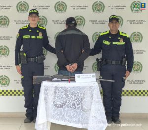 En la imagen se observa el procesado de espaldas, en medio de dos integrantes de la Policía Nacional. También se evidencia la escopeta que fue incautada durante el procedimiento de captura.