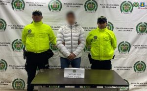 En la imagen se observa a una persona capturada, ubicada en el centro, con el rostro desenfocado y las manos juntas al frente, custodiada por dos uniformados de la Policía Nacional de Colombia. Los policías visten chaquetas institucionales de alta visibilidad color verde y gorras con el distintivo “Policía”.Al fondo hay un pendón oficial repetido con el escudo y el nombre de la Policía Nacional de Colombia, así como el logo de la Fiscalía General de la Nación en la parte superior derecha. Frente a los tres aparece una mesa sobre la cual se encuentra un aviso que dice “Captura por orden judicial”. En la esquina inferior derecha de la imagen se lee la leyenda: “Insumo de comunicación con fines periodísticos”. La escena corresponde a un registro oficial de una captura realizada por las autoridades.