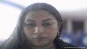 En la imagen se observa una mujer en primer plano, aparentemente captada a través de una cámara o videollamada.Tiene cabello largo y oscuro, peinado hacia atrás, y lleva aretes tipo argolla.  Su expresión es seria o concentrada, con la mirada dirigida ligeramente hacia abajo, como si estuviera leyendo o escuchando algo.  Viste una prenda oscura, posiblemente una chaqueta o blazer.  El fondo está desenfocado, pero parece un espacio interior, posiblemente una oficina o sala. Se distingue una figura al fondo con ropa azul, lo que sugiere que hay más personas en el lugar.  En la esquina inferior derecha aparece el texto: “Insumo de comunicación con fines periodísticos”.