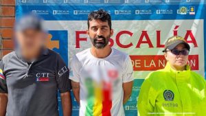 El procesado se encuentra esposado y en custodia por un servidor del Cuerpo Técnico de Investigación (CTI) y una uniformada de la Policía. Detrás de él hay un backing de la Fiscalía. 