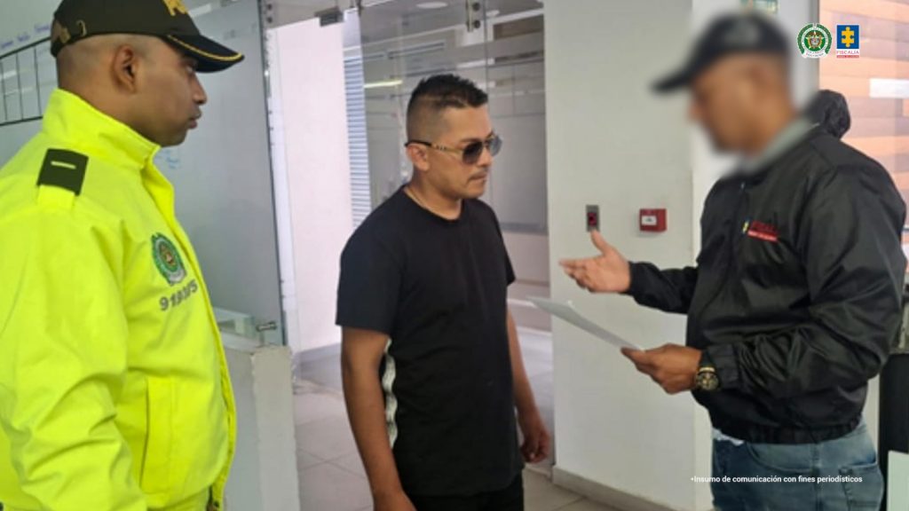 La fotografía fue tomada dentro de unas oficinas y se aprecia de pie y de perfil a William Andrés Rayo Jiménez. Viste camiseta de manga corta, de color negro y pantalón negro. Frente a él hay un servidor del CTI quien está en acción de lectura de un documento. Al extremo derecho del capturado hay un uniformado de la Policía Nacional.
