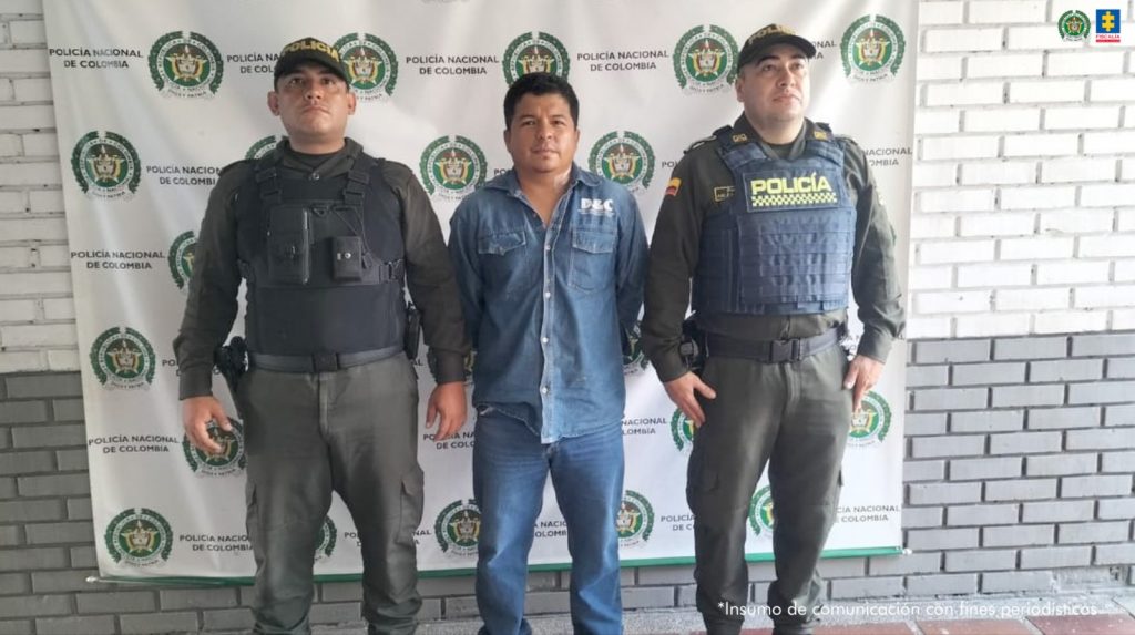 En la fotografía se aprecia de pie y con los brazos atrás a William Marino Mambuscay Salinas. Viste con camisa de manga larga, azul y de botones; usa jean azul. A sus extremos hay uniformados de la Policía Nacional. Detrás de ellos hay un pendón institucional de la Policía.