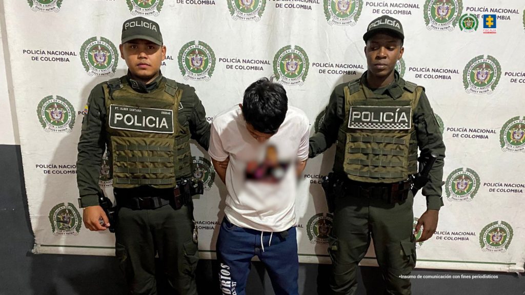 Se visualiza al capturado junto a dos uniformados de la Policía Nacional. Detrás de ellos el banner que identifica a la Policía Nacional
