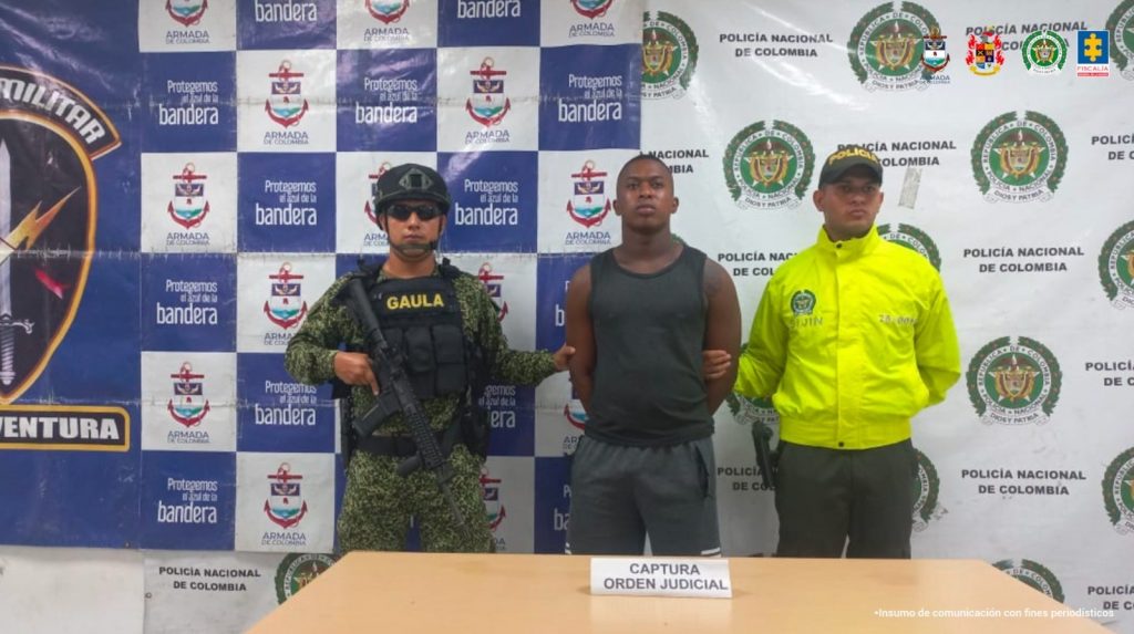 capturado de frente custodiado por dos policías. Detrás banner de la Policía, la Armada y el Ejército Nacional.