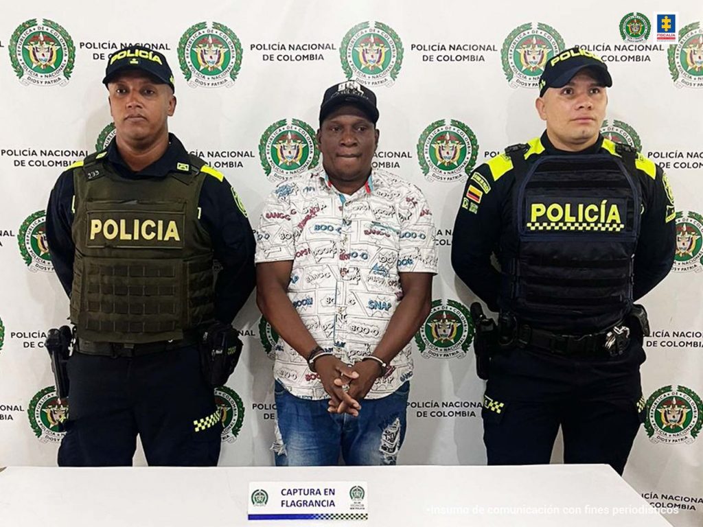 En la fotografía se aprecia de pie, con los brazos adelante y esposado a José Willinton Ortiz Castillo. Viste camisa blanca con estampados de color negro, azul y rojo; gorra negra y jean azul desgastado. A lado y lado de él hay uniformados de la Policía Nacional. Delante de ellos hay un escritorio blanco y sobre él un letrero con logos de la Policía que dice: ‘Captura en flagrancia’. Detrás de ellos hay un pendón institucional de la Policía.