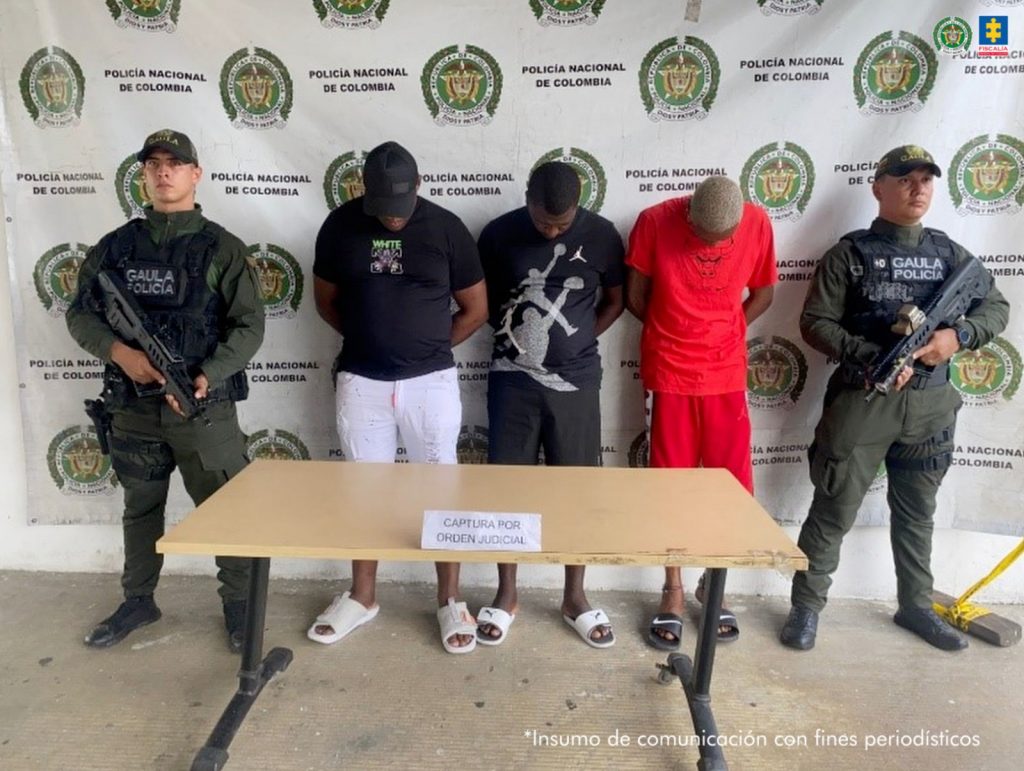 En la fotografía aparecen tres de los cuatro presuntos implicados. Uno, viste camiseta negra, de manga corta, gorra negra y bermuda blanca; otro viste camiseta de manga corta y bermuda, ambas de color negro; un tercero, viste camiseta de manga corta y bermuda de color rojo; su cabello esta decolorado. Los tres tienen inclinada la cabeza. A sus extremos hay uniformados de la Policía Nacional. Delante de ellos hay una mesa y sobre ella hay un letrero que dice: ‘Capturas por orden judicial’. Detrás de ellos hay un pendón institucional de la Policía.