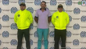  En la fotografía aparece un hombre capturado, acompañado de dos uniformados de la Policía Nacional. En la parte posterior un banner con logos de la entidad. 
