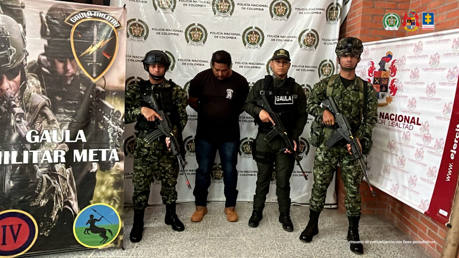 capturado de frente custodiado por dos militares y un policía. Detrás banner de grupo Gaula, Policía y Ejército Nacional.