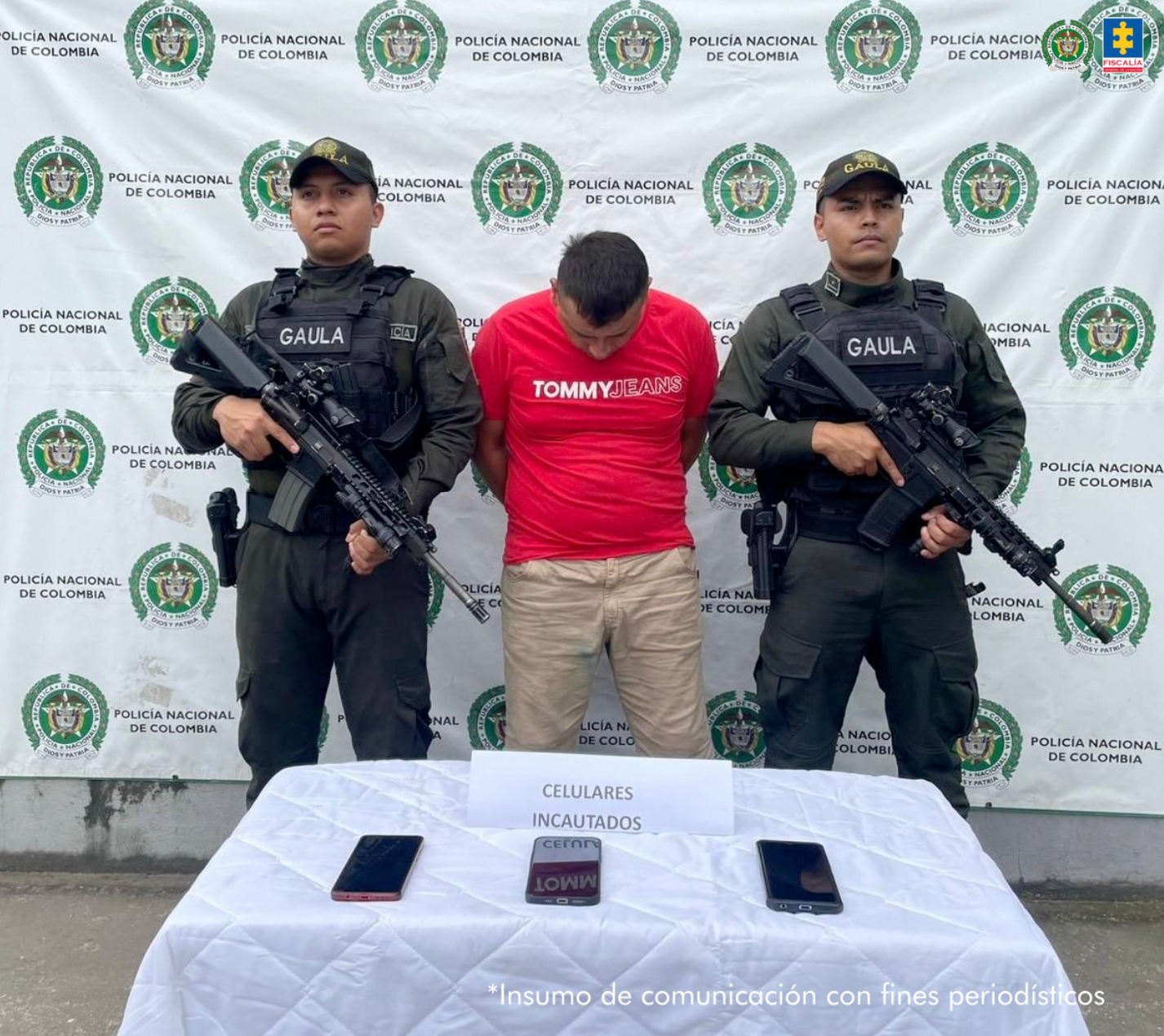 capturado de frente custodiado por dos policías del grupo Gaula. al frente de ellos una mesa con material incautado. Detrás banner de la Policía Nacional.