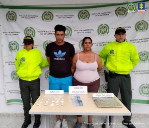 En la fotografía se aprecian de pie y con los brazos atrás a Wuilimar Mairobis Mambel Rodríguez y Abraham Antonio Díaz Carreño. El hombre viste camiseta negra de manga corta ccon logo de color azul; bermuda azul y zapatillas blancas. La mujer viste una blusa de tiras de color rosado y pantalones de color café. A sus extremos hay uniformados de la Policía Nacional. Delante de ellos hay una mesa color marrón y sobre ella varios elementos como papeletas con contenido blanco, billetes y una bolsa plástica con contenido verdoso. Detrás de ellos hay un pendón institucional de la Policía.