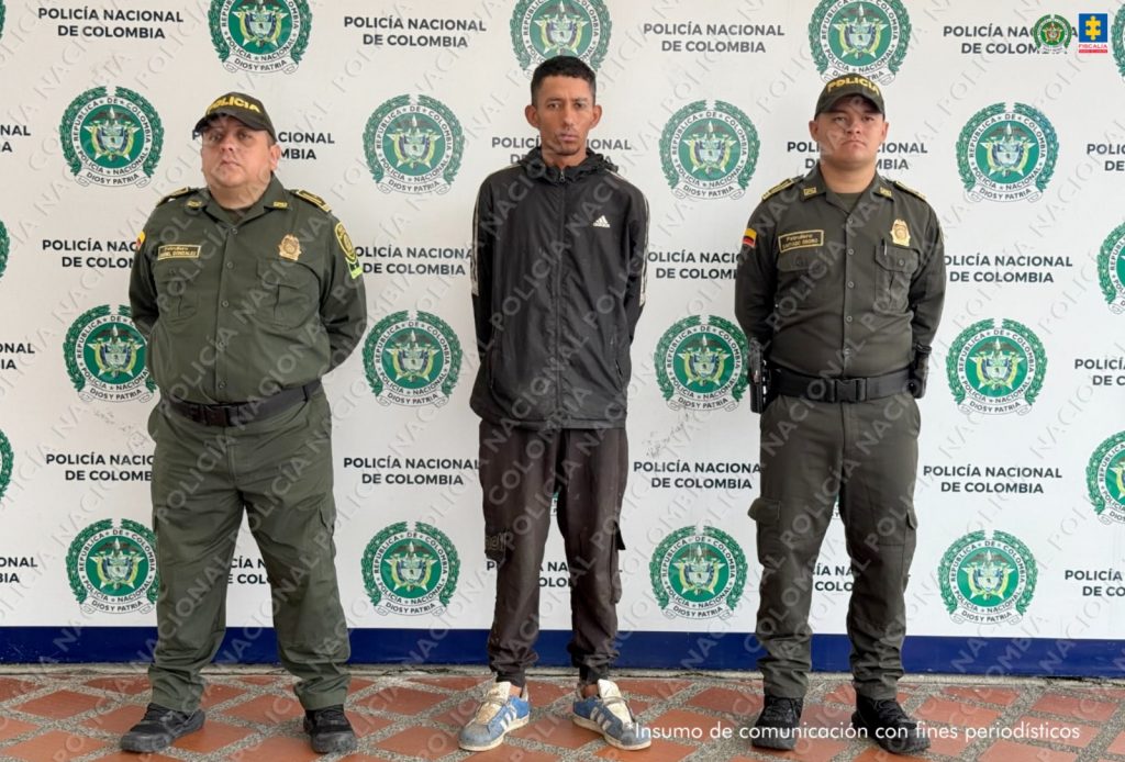 Se observa al capturado de frente y a cada costado un funcionario de Policía nacional. Atrás, un pendón de Policía Nacional