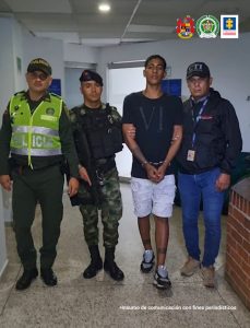 En la foto aparece en un pasillo, en acción de desplazamiento, con los brazos adelante y esposado, Yan Alexánder Muñoz Gómez. Viste Camiseta negra de manga corta, pantaloneta blanca y zapatillas negras con blanco. Al lado derecho de él hay dos uniformados de la Policía y del Ejército. Al lado izquierdo hay un servidor del CTI de la Fiscalía.