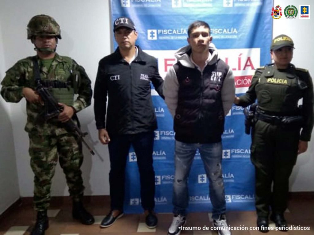 En la foto aparece Yan Carlos Fernández Astudillo, con los brazos atrás, vestido con una chaqueta oscura con mangas grises, jean azul y zapatillas blancas. Al lado derecho de él hay un servidor del CTI de la Fiscalía y un militar. A su izquierda una uniformada de la Policía Nacional. Detrás de ellos hay un pendón institucional de la Fiscalía.