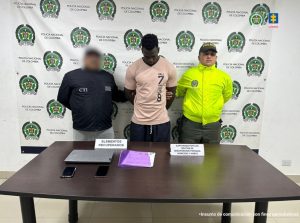 En la fotografía se aprecia de pie y con los brazos atrás a Yanier Hurtado Asprilla, alias Max. Viste camiseta de color curuba, de manga corta y con estampadao vertical de letras de color negro al lado izquierdo; pantalón negro. Al lado derecho de él hay un servidor del CTI de la Fiscalía y al lado derecho un uniformado de la Policía Nacional. Delante de ellos hay una mesa de color marrón y sobre ella hay un computador portátil, dos celulares y una carperta plástica de color púrpura con documentos. Detrás de ellos hay un pendón institucional de la Policía.