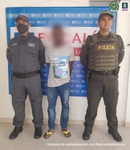 capturado de frente esposado manos atrás, custodiado por investigador del CTI de la Fiscalía y un uniformado de la Policía. Detrás banner de la Fiscalía Nacional.