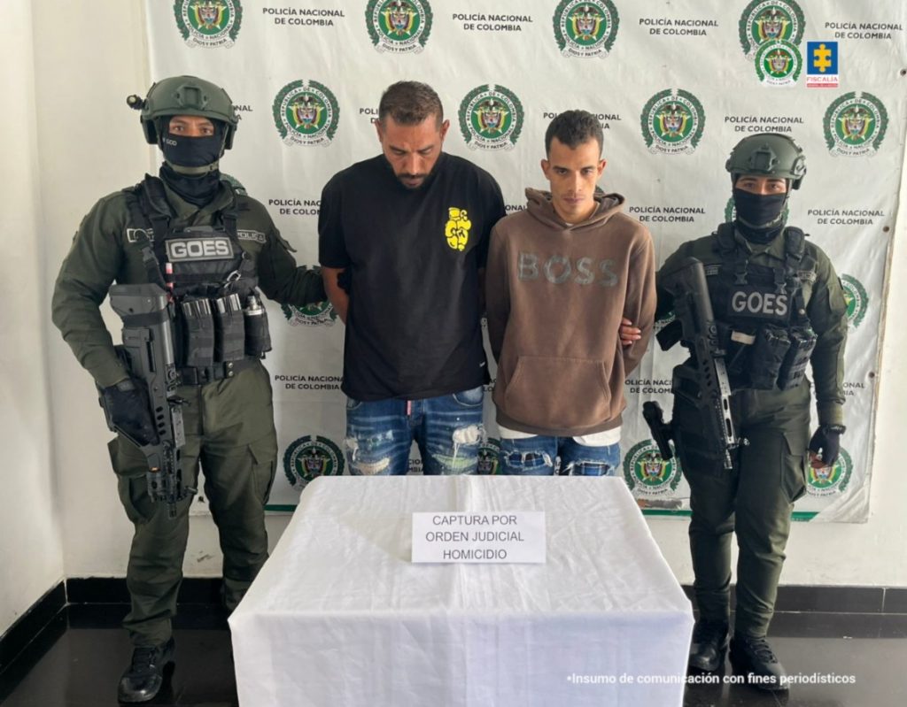 Se observan los dos capturados de frente y a cada costado un servidor de Policía Nacional que los custodia. Atrás un pendón de Policía Nacional
