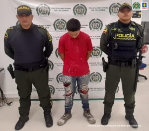 capturado de frente cabeza agachada y esposado manos atrás, custodiado por dos uniformados de la Policía Nacional, detrás banner de la Policía Nacional.