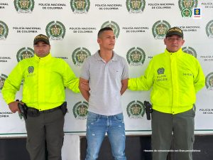 En la fotografía aparece un hombre capturado, acompañado de dos uniformados de la Policía. En la parte posterior un banner con logos de la entidad.