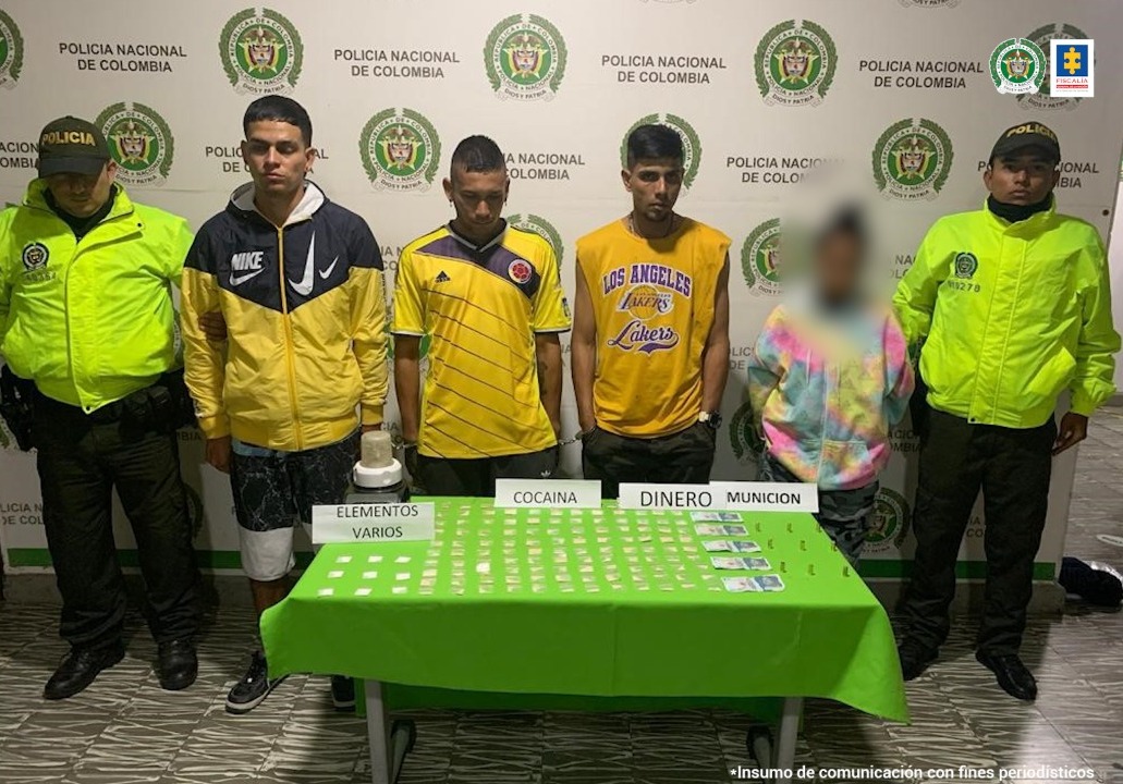 En la foto aparecen los presuntos implicados, acompañados por personal de la Policía Nacional. Delante de ellos hay una mesa con mantel verde y sobre ellas varios elementos y letreros que dicen ‘cocaína’, ‘munición’, ‘dinero’ y ‘elementos varios’. Detrás de ellos hay un pendón institucional de la Policía.