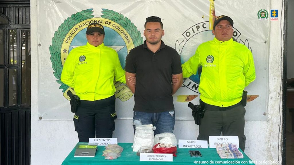 Se visualiza al capturado junto a dos uniformados de la Policía Nacional. Frente a ellos una mesa con los elementos incautados (sustancias alucinógenas, gramera, bolsas herméticas y dinero) Detrás de ellos el banner que identifica a la Policía Nacional.