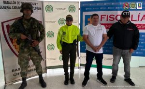 está el capturado de frente esposado manos adelante, custodiado por investigador de CTI Fiscalía, policía y soldado. Detrás banner de Fiscalía, Policía y Ejército Nacional.