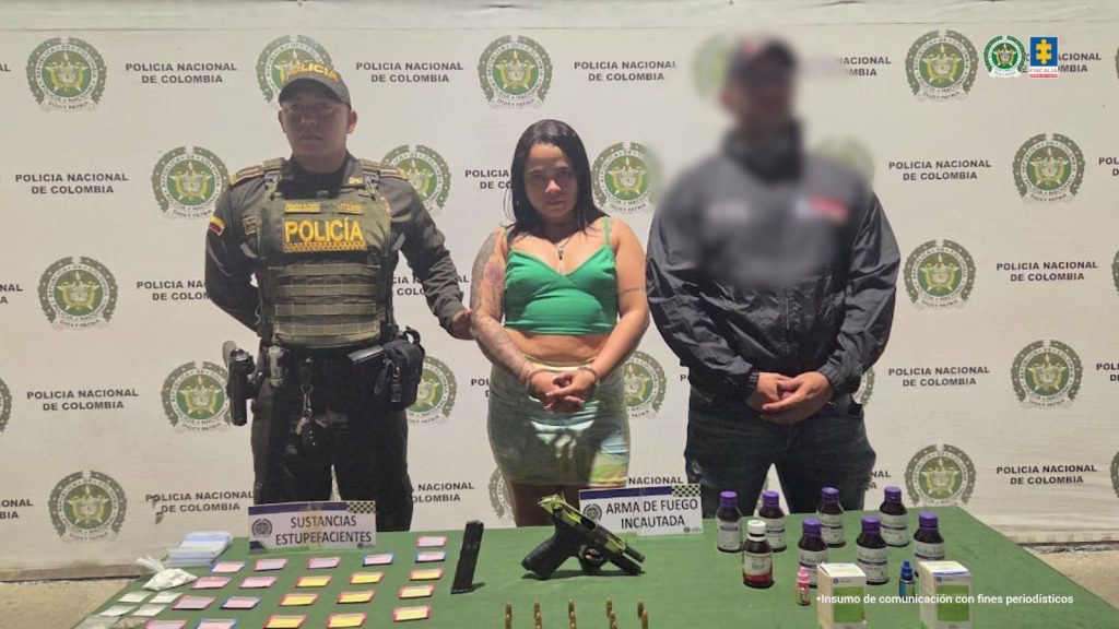 En la fotografía aparece de pie, con los brazos adelante y esposada Yerly Marcela López Giraldo. Viste con blusa de tiras, de color verde y ombliguera; falda de jean azul claro. A sus extremos hay uniformados de la Policía Nacional. Delante de ellos hay una superficie de color verde y sobre ella se aprecian bolsas plásticas transparentes, un arma de fuego, un proveedor, munición, un celular y frascos. Detrás de la judicializada hay un pendón institucional de la Policía.