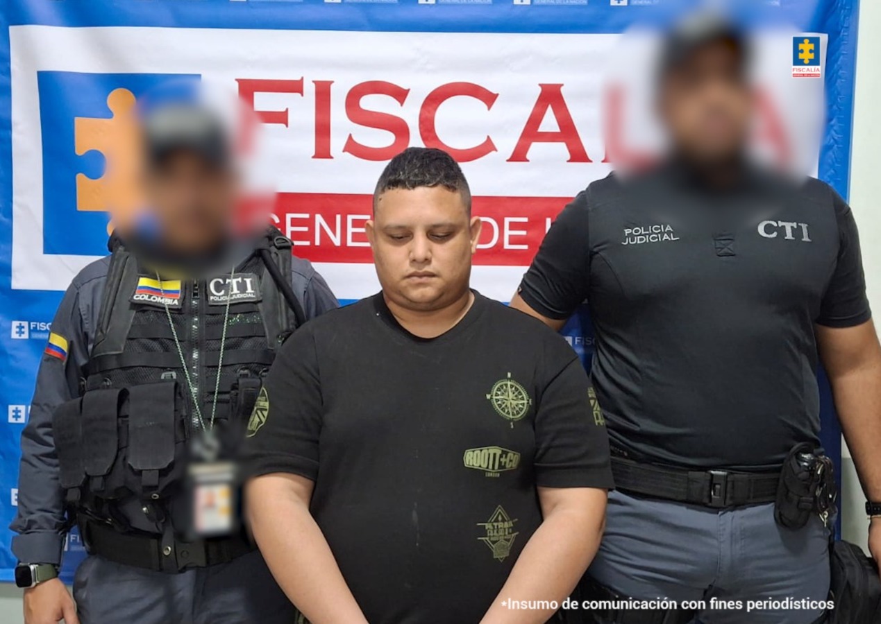 El hombre aparece de pie, de medio cuerpo, con las manos esposadas adelante. Está rodeado de miembros del CTI de la Fiscalía. Hay un backing de la institución al fondo. 