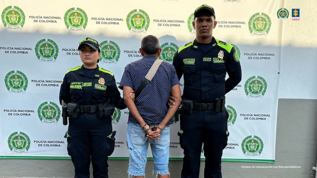  capturado de espaldas custodiado por dos policías. Detrás banner de la Policía Nacional.