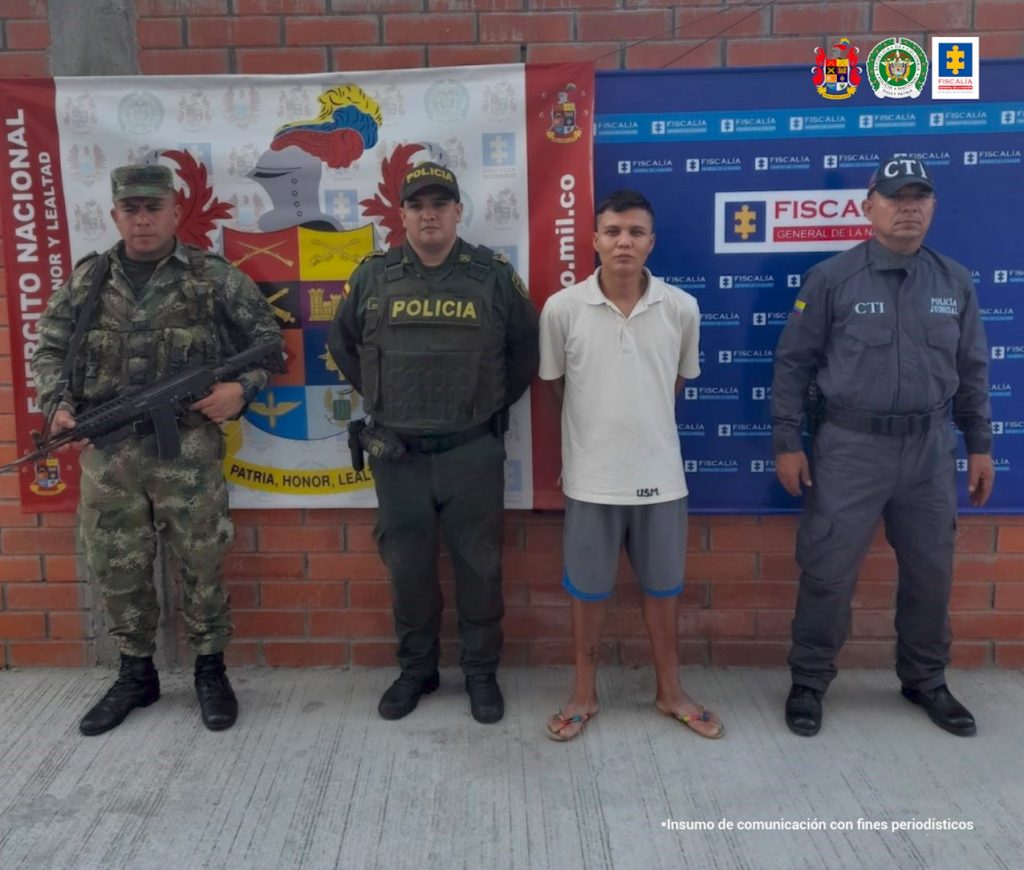 En la foto aparece de pie y con los brazos atrás Yohan Sebastián Ninco Vargas. Viste camisa blanca, bermudas de color gris con dobladillo azul y pantuflas. Al lado derecho hay uniformados de la Policía Nacional y del Ejército Nacional. A la izquierda hay un servidor del CTI de la Fiscalía. Detrás de ellos hay dos pendones institucionales del Ejército y la Fiscalía.