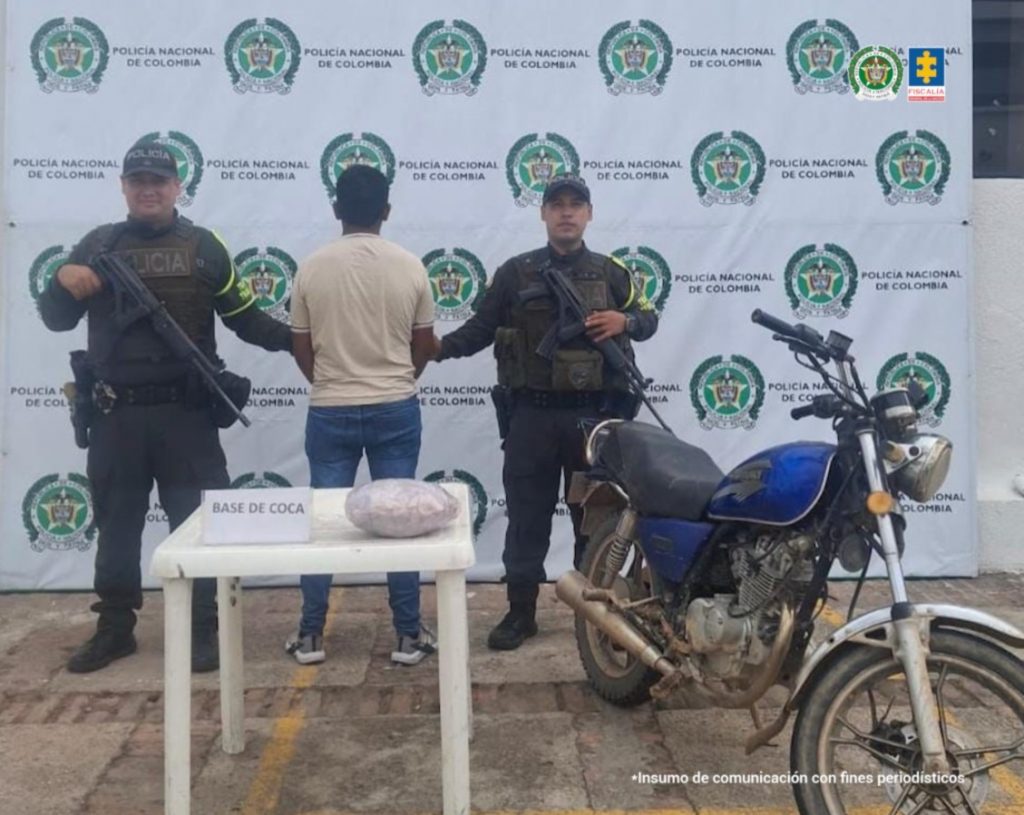 Se observa al capturado de espaldas junto a dos uniformados de la Policía Nacional. Frente a ellos una mesa con la sustancia estupefaciente incautada y la motocicleta utilizada para transportar la sustancia. Detrás de ellos el banner que identifica a la Policía Nacional.